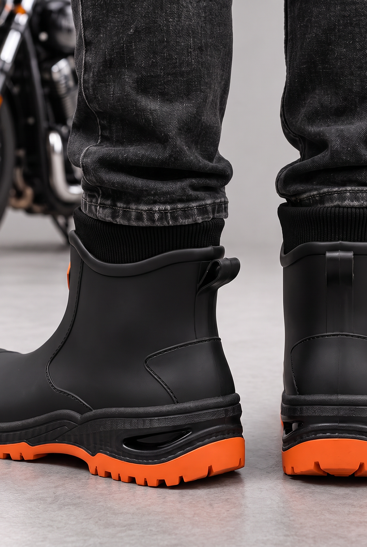 Black ankle rain boots with orange lugged soles worn with dark jeans, back view with pull tabs.