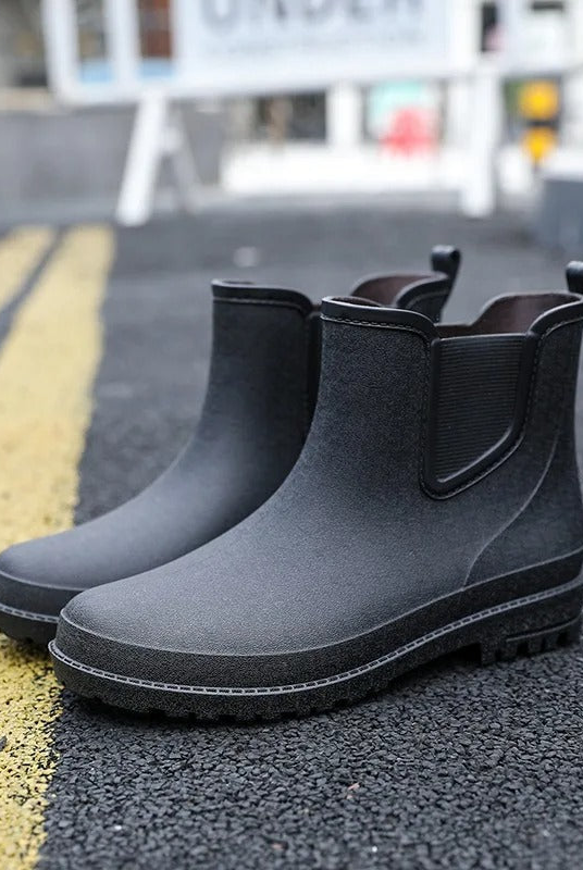 A pair of black ankle high Chelsea style rubber rain boots with elastic side panels and pull tabs, standing on an asphalt road beside yellow road markings, with a blurred street background.