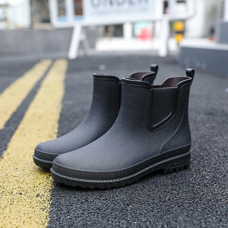 A pair of black ankle high Chelsea style rubber rain boots with elastic side panels and pull tabs, standing on an asphalt road beside yellow road markings, with a blurred street background.