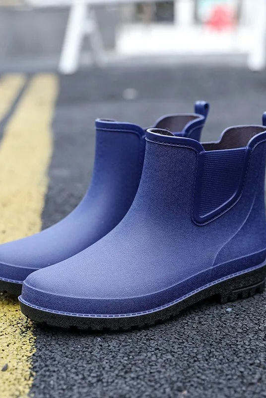 A pair of navy blue rubber Chelsea style ankle boots with black soles and elastic side panels, standing on an asphalt road beside yellow road markings.
