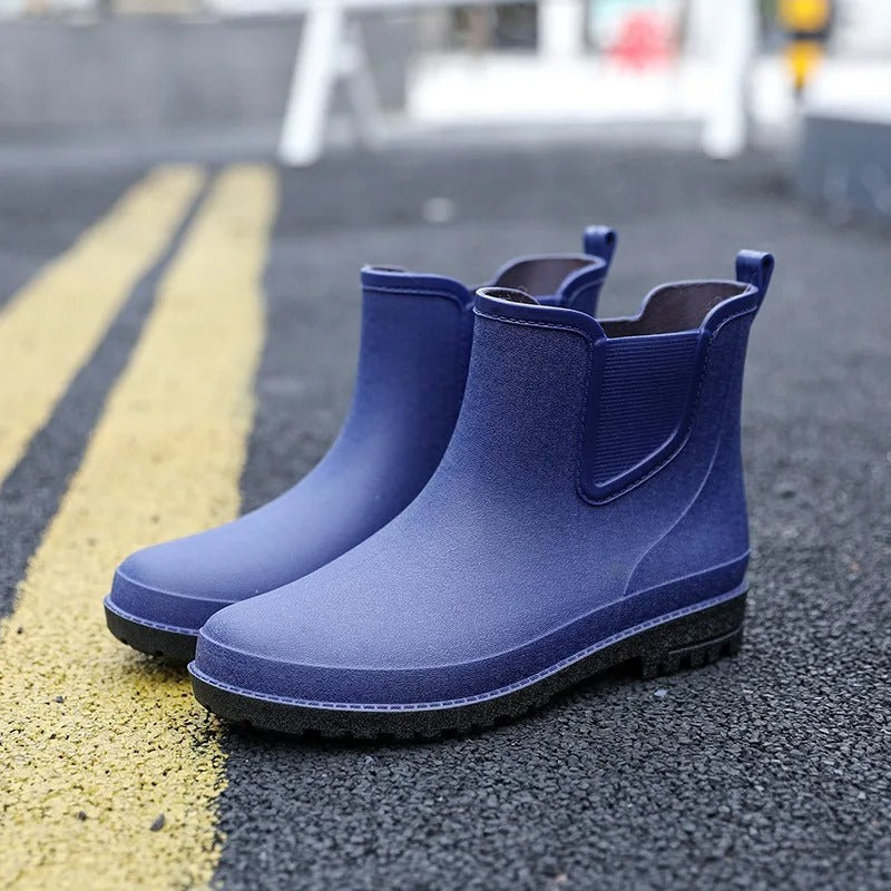 A pair of navy blue rubber Chelsea style ankle boots with black soles and elastic side panels, standing on an asphalt road beside yellow road markings.