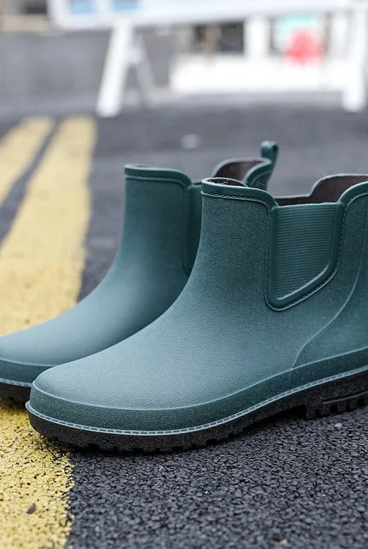 A pair of dark green rubber Chelsea style ankle boots with elastic side panels, pull tabs and black soles, standing on an asphalt road beside yellow lane markings.