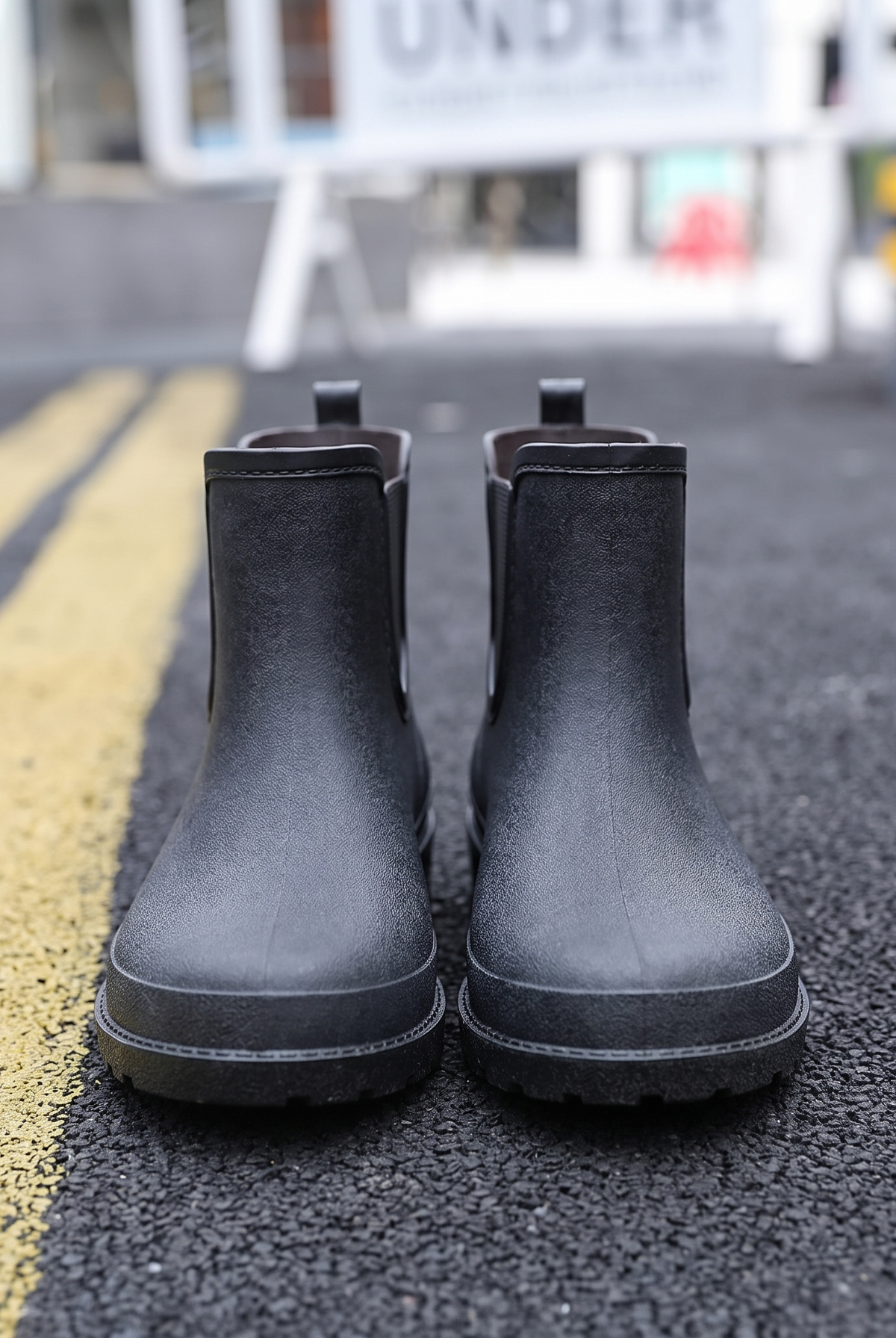 A pair of black rubber Chelsea style rain boots with elastic side panels and pull tabs standing on a street with yellow road markings, front view