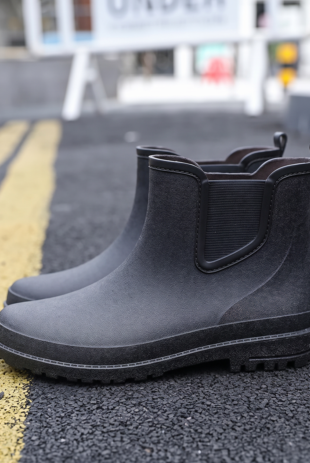 A pair of black rubber Chelsea style rain boots with elastic side panels and pull tabs standing on a street with yellow road markings, side view