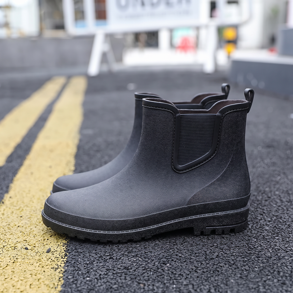 A pair of black rubber Chelsea style rain boots with elastic side panels and pull tabs standing on a street with yellow road markings, side view
