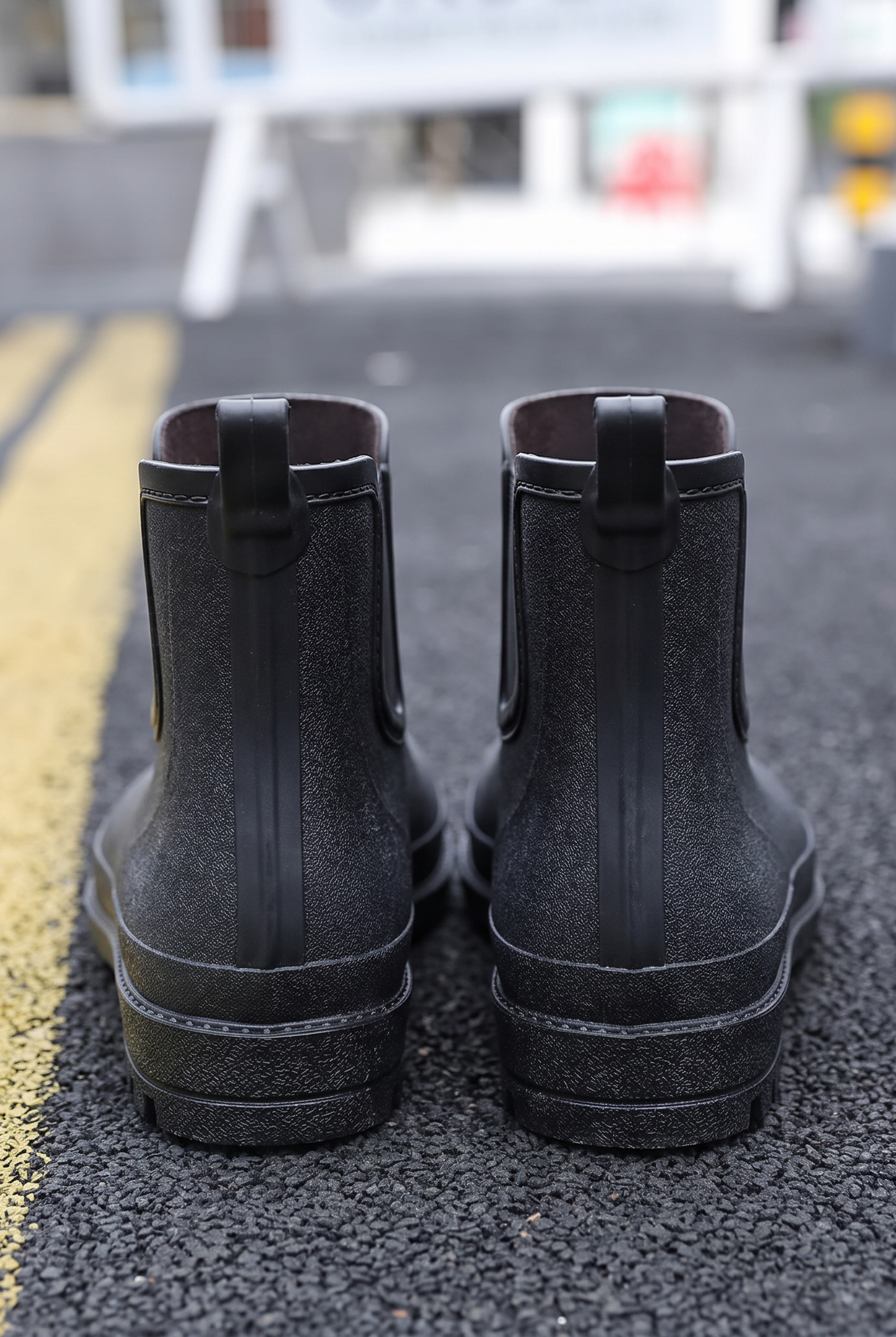 A pair of black rubber Chelsea style rain boots with elastic side panels and pull tabs standing on a street with yellow road markings, back view