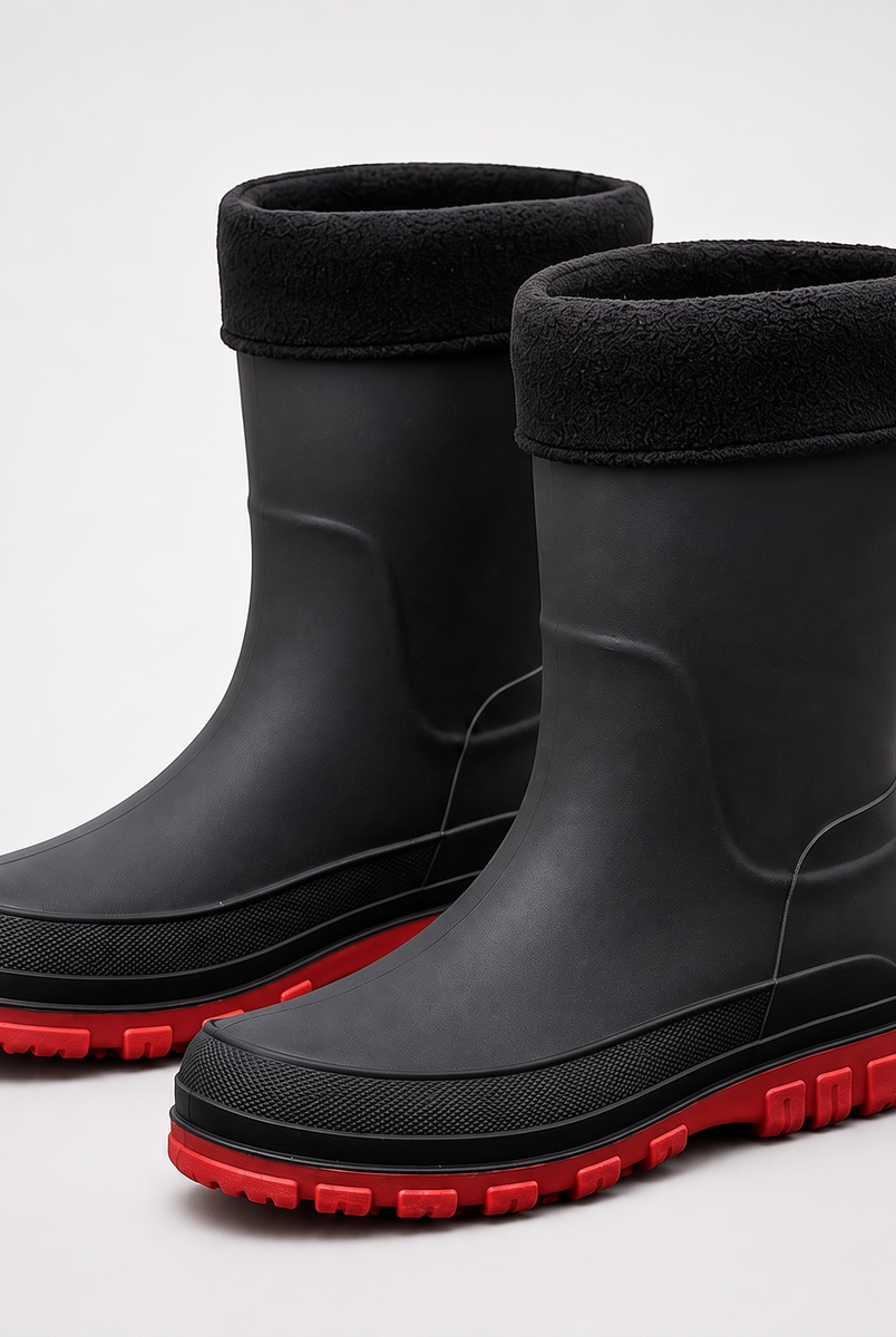 A pair of black insulated rain boots with a black fleece cuff and a red lugged sole, shown on a plain grey studio background.
