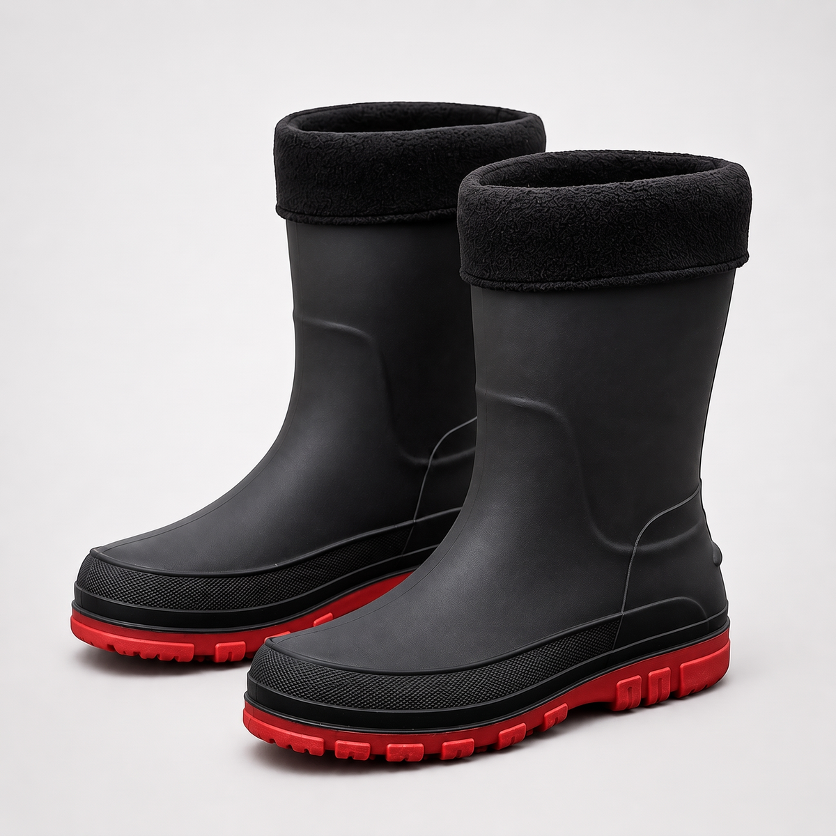 A pair of black insulated rain boots with a black fleece cuff and a red lugged sole, shown on a plain grey studio background.