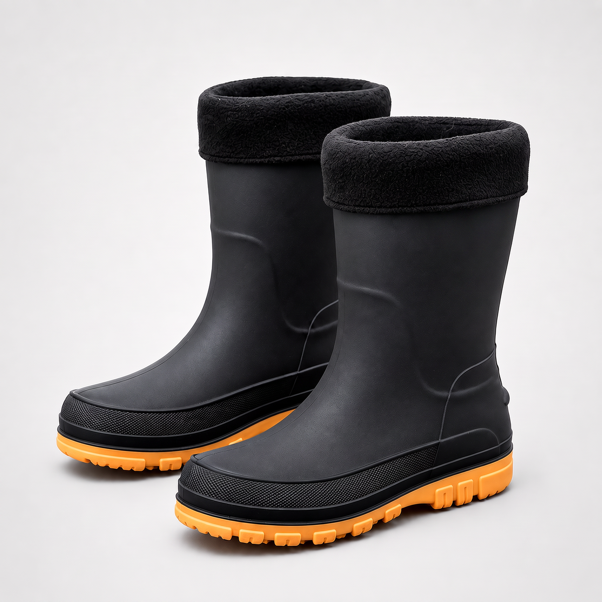 Studio three quarter view of a pair of black rubber rain boots with black fleece cuffs, a textured black welt band and chunky orange soles.