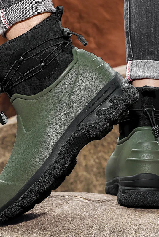 A person in rolled grey jeans stands on rocks outdoors wearing a pair of olive green rubber ankle boots with black neoprene cuffs, elastic toggle laces and thick black lug soles.