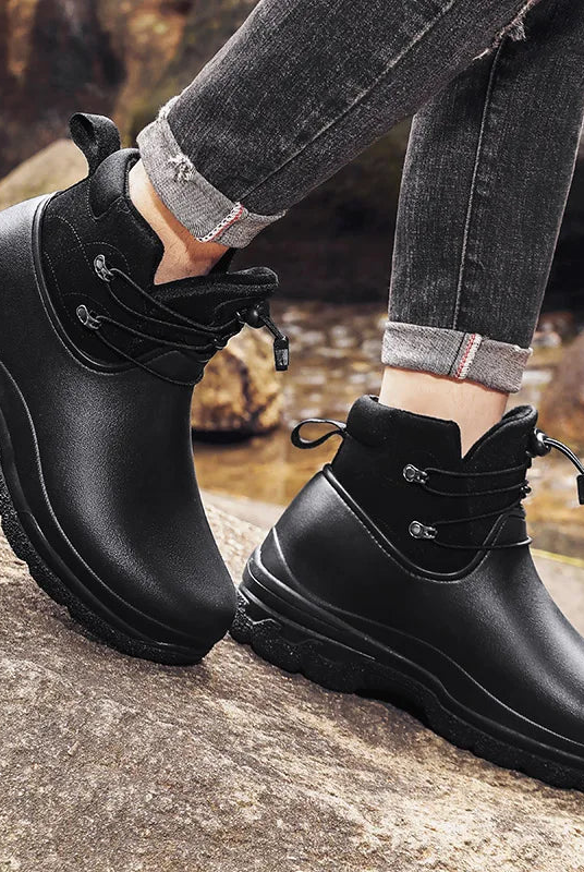 A person in cuffed grey jeans wears a pair of black ankle rain boots with a suede-look collar and bungee lacing, standing on rocks outdoors beside a stream.