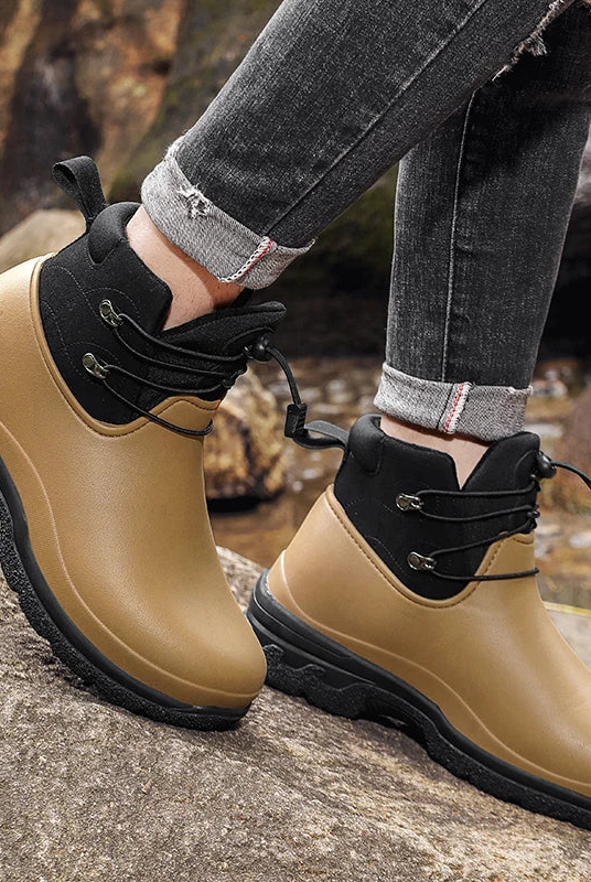 A person in rolled grey jeans wears a pair of tan rubber ankle boots with a black padded collar, elastic drawstring laces and chunky black lug soles, standing on rocks outdoors near a stream.