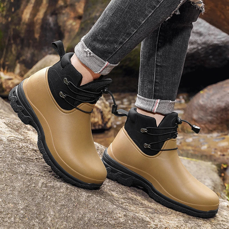A person in rolled grey jeans wears a pair of tan rubber ankle boots with a black padded collar, elastic drawstring laces and chunky black lug soles, standing on rocks outdoors near a stream.