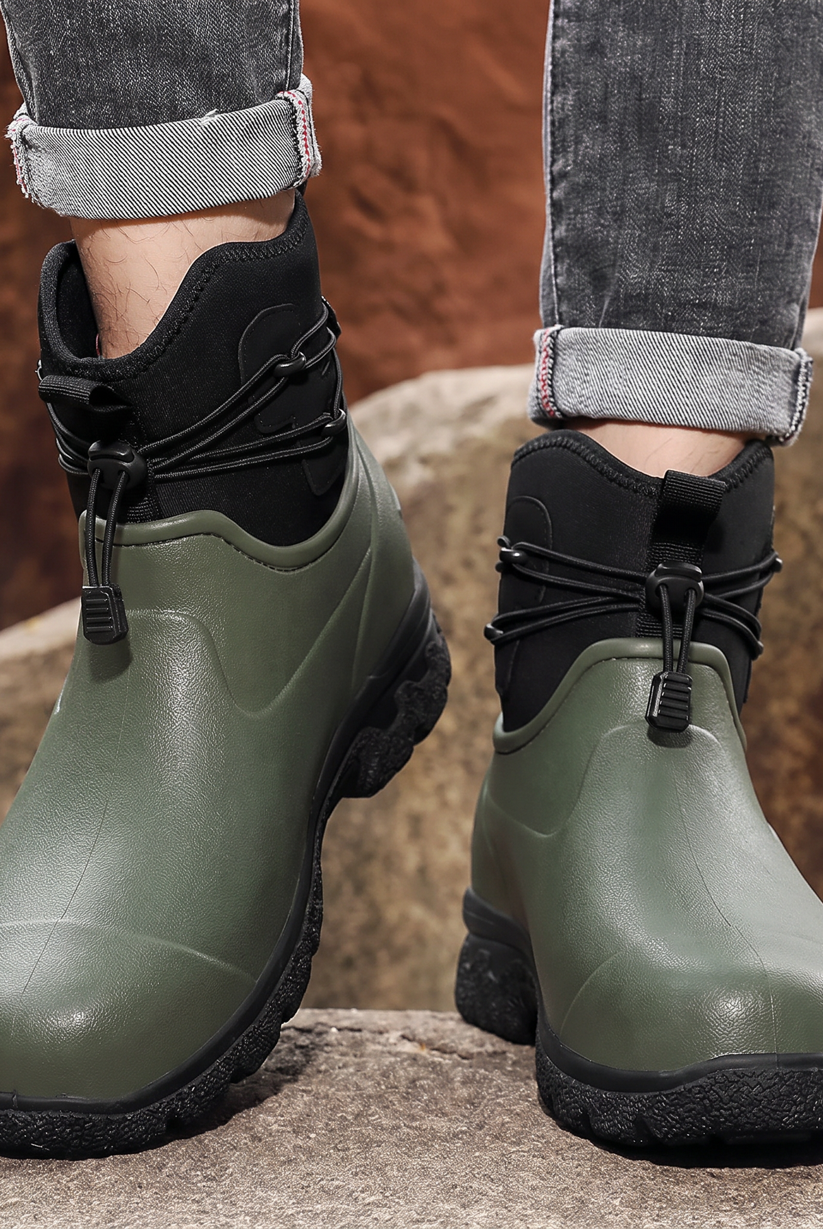A person in rolled grey jeans stands on rocks outdoors wearing a pair of olive green rubber ankle boots with black neoprene cuffs, elastic toggle laces and thick black lug soles, front view