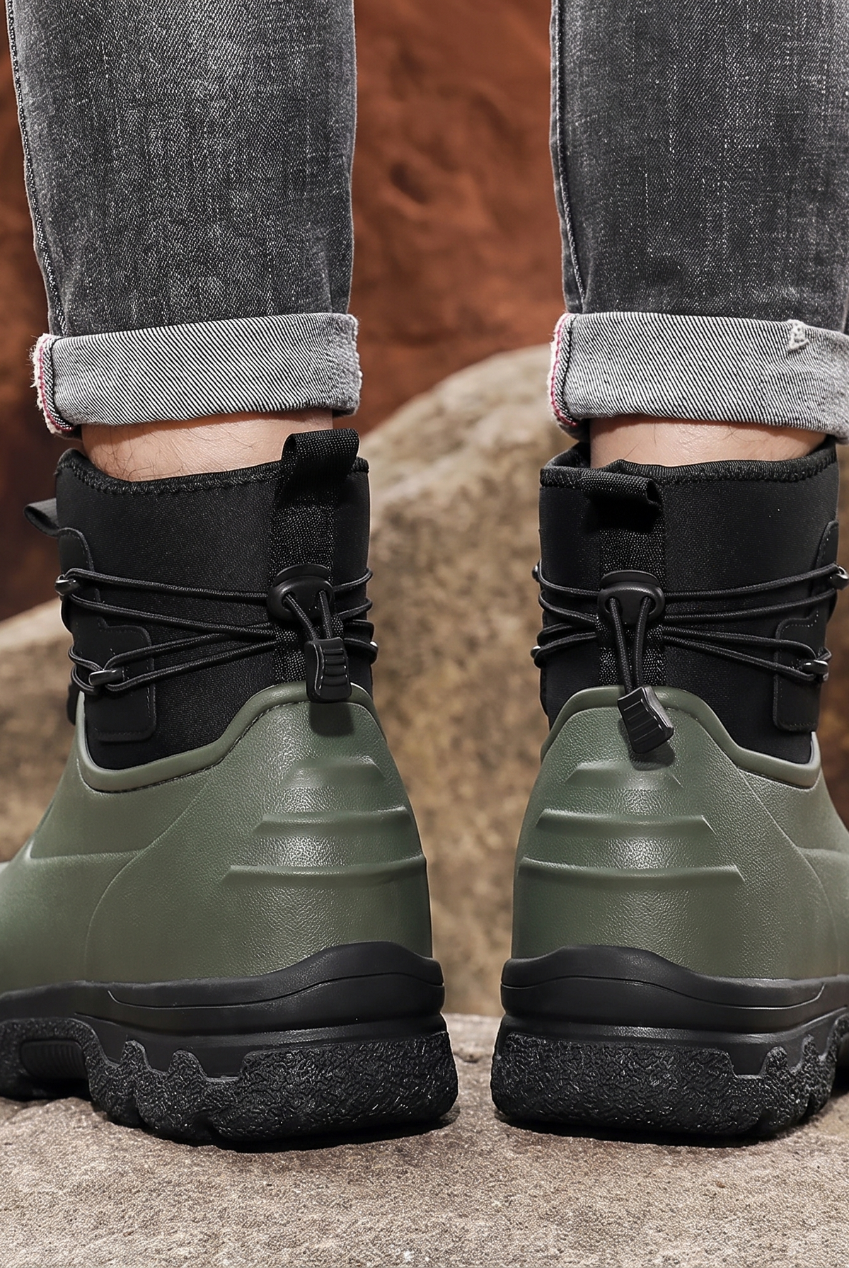 A person in rolled grey jeans stands on rocks outdoors wearing a pair of olive green rubber ankle boots with black neoprene cuffs, elastic toggle laces and thick black lug soles, back view