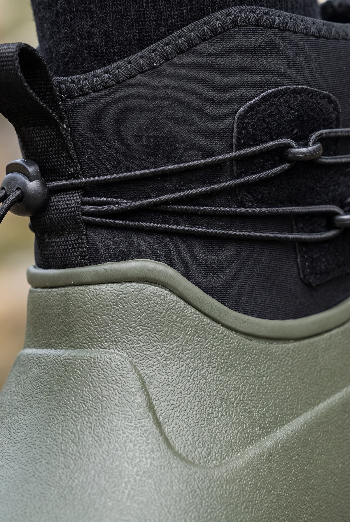 Olive green rubber ankle rain boot with a black neoprene collar, bungee drawstring lacing and a black lugged sole, shown in three quarter view on a white background, close-up view