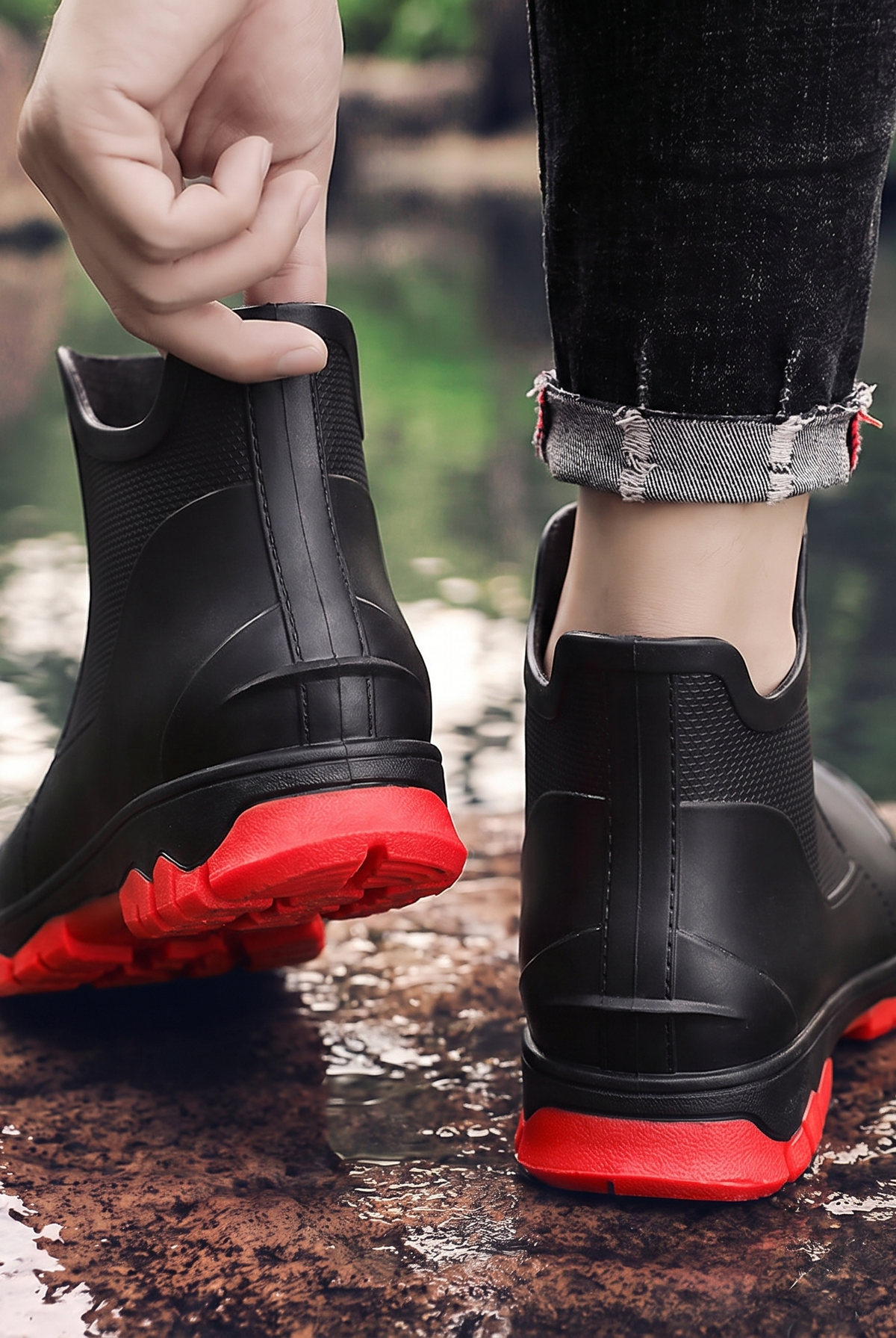 A pair of black ankle rain boots with red soles, one worn on a foot and one held by a hand, photographed outdoors near water, back view