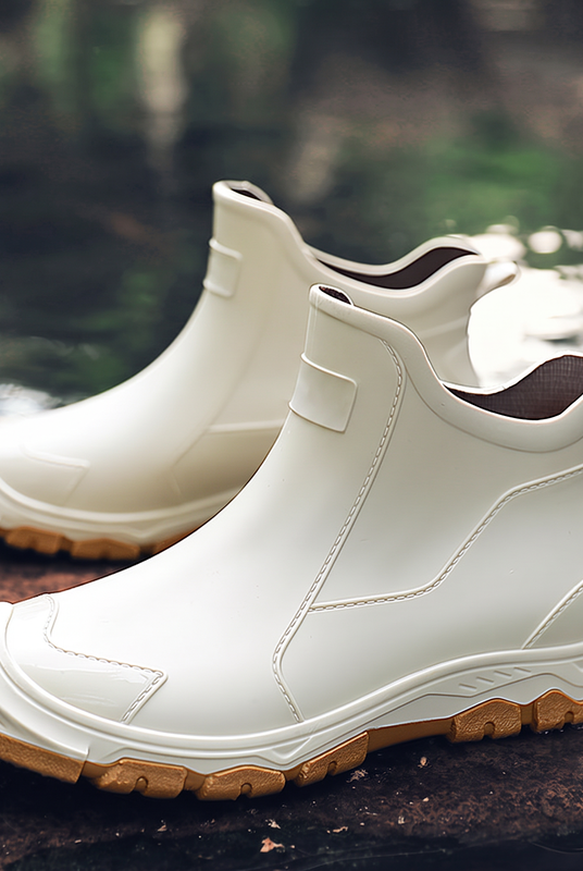 A pair of white ankle-high rubber boots with tan soles displayed outdoors on a wet rock near water.