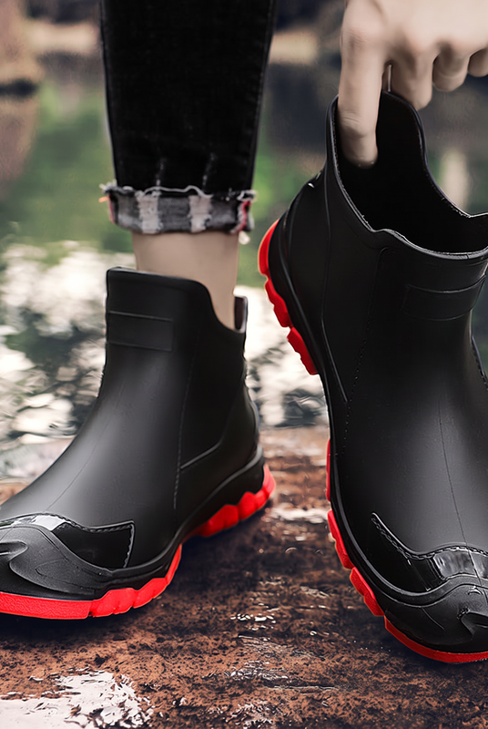 A pair of black ankle rain boots with red soles, one worn on a foot and one held by a hand, photographed outdoors near water.