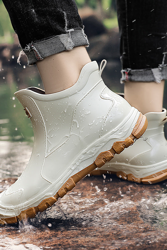 A person wearing cream colored ankle rain boots with tan textured soles stands on wet rocks near water