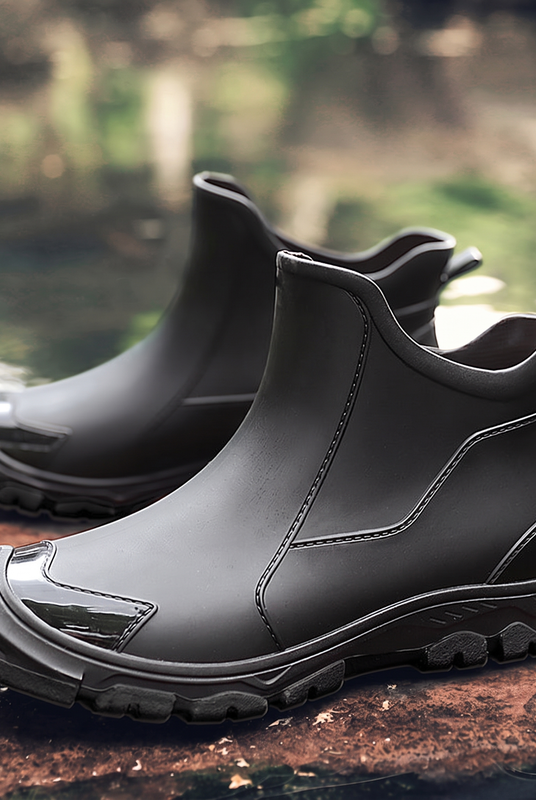 A pair of black rubber ankle boots with a side logo patch, shown on a rock near water.