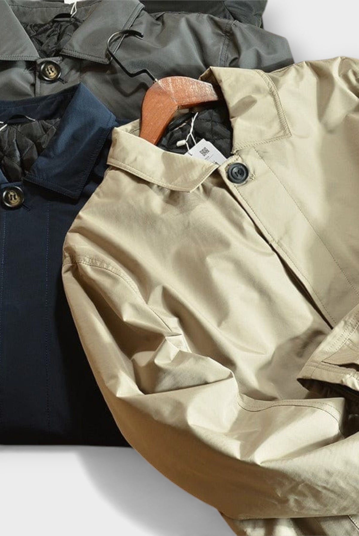 Beige, navy, and olive button-front rain macs stacked flat, folded overlapping | Chaps London