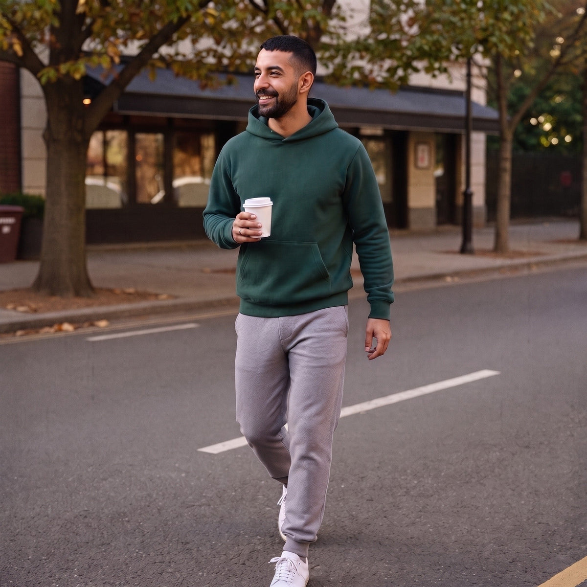 Man wearing dark green pullover hoodie with grey sweatpants, walking outdoors holding coffee | Chaps London