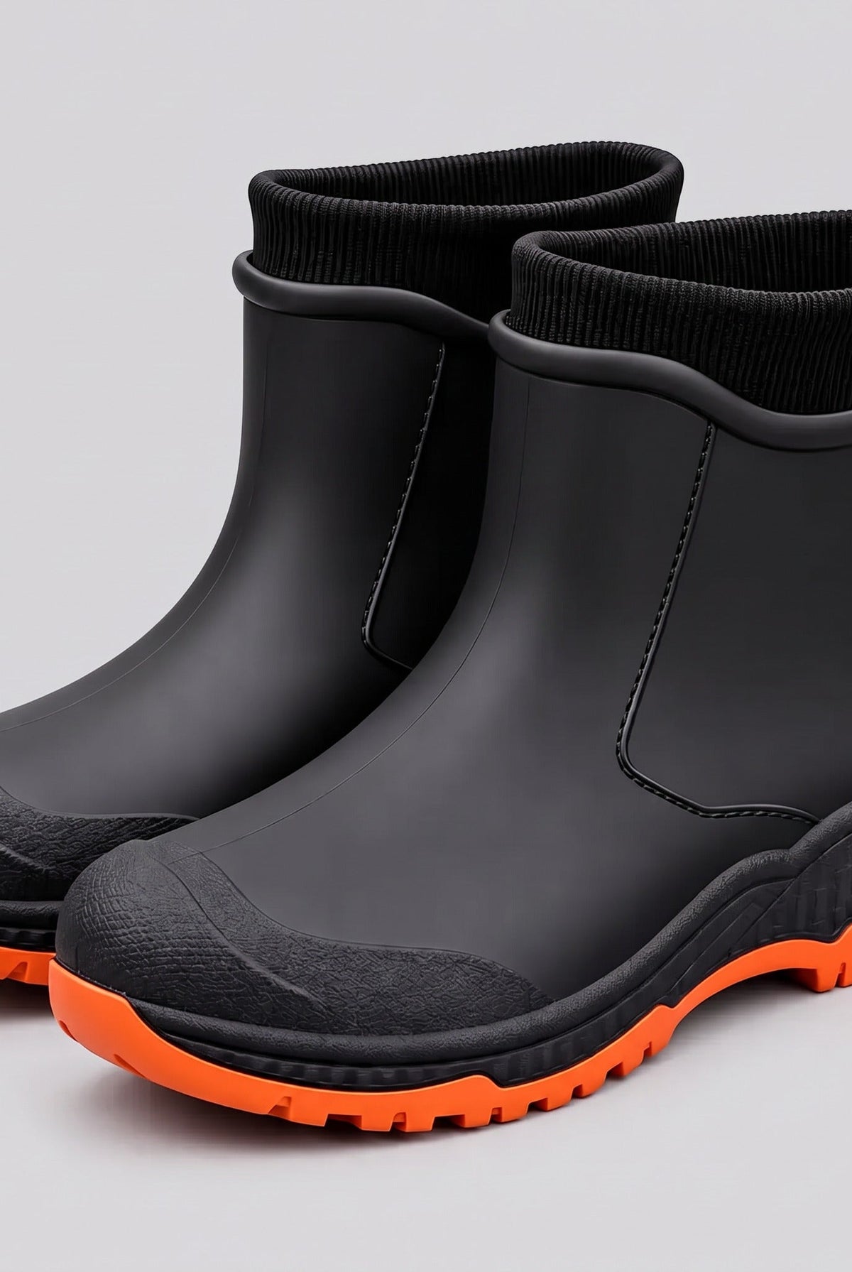 Black ankle rain boots with orange lugged soles and black ribbed knit cuffs, three quarter view on a grey studio background.