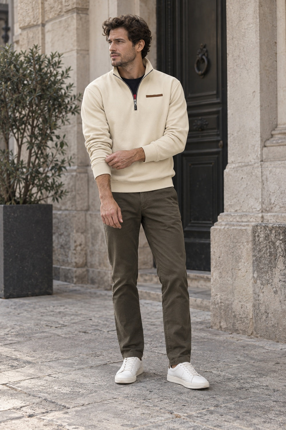 Full-length view of a man in a beige quarter zip sweatshirt with olive chinos and white trainers by a stone doorway | Chaps London