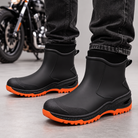 Black ankle rain boots with orange lugged soles worn with dark jeans, motorcycle in the background.