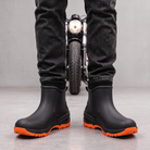 Black ankle rain boots with orange lugged soles worn with dark jeans, front view.