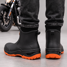 Black ankle rain boots with orange lugged soles worn with dark jeans, back view with pull tabs.