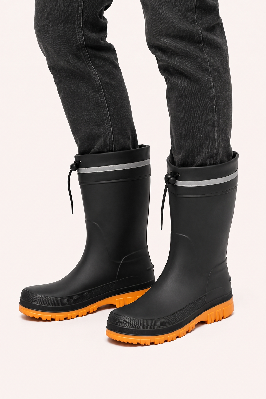 Studio photo of a person in dark grey jeans wearing black mid calf rain boots with orange lugged soles, a drawstring cuff and a reflective grey band.