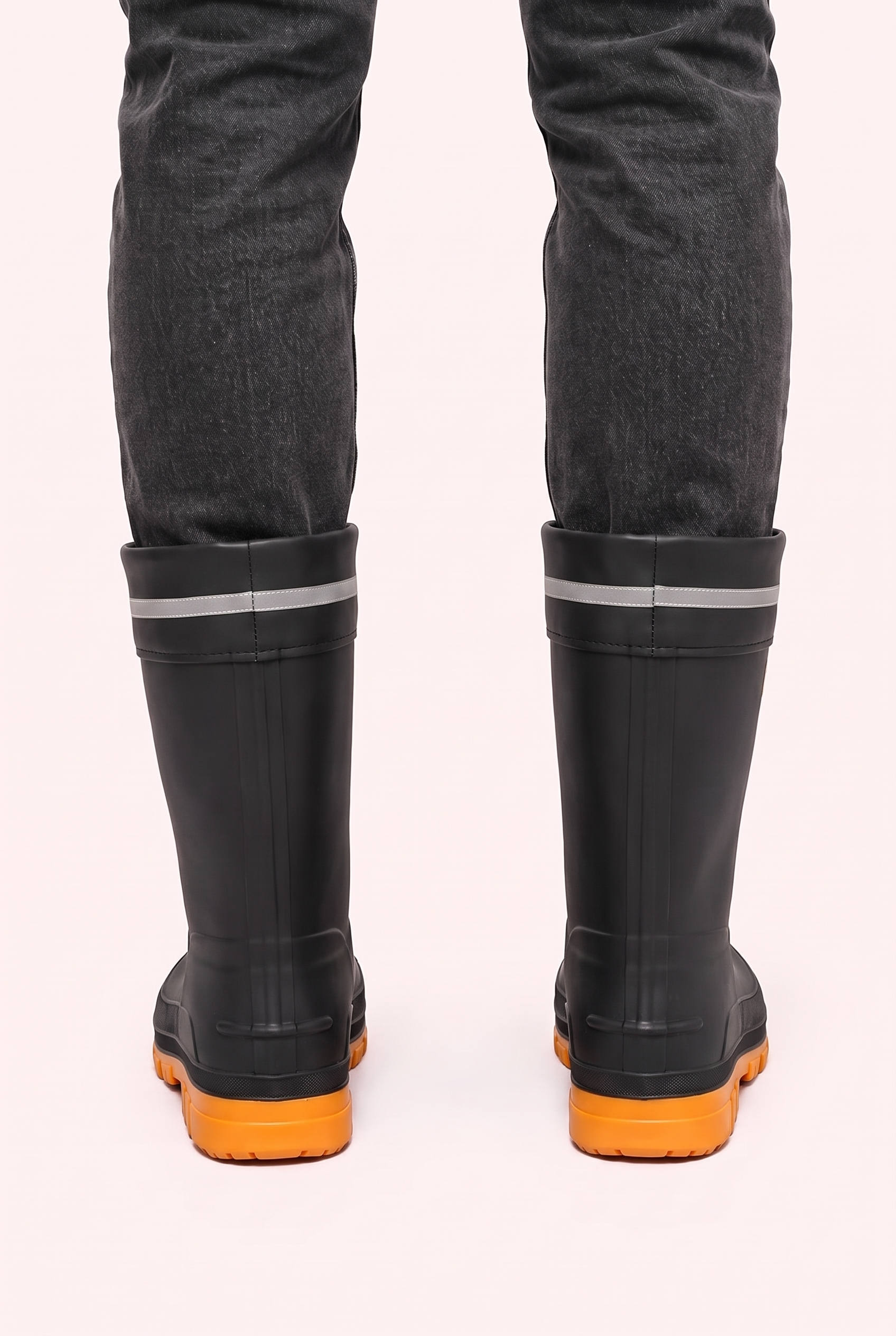 Studio photo of a person in dark grey jeans wearing black mid calf rain boots with orange lugged soles, a drawstring cuff and a reflective grey band, back view