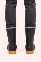 Studio photo of a person in dark grey jeans wearing black mid calf rain boots with orange lugged soles, a drawstring cuff and a reflective grey band, back view