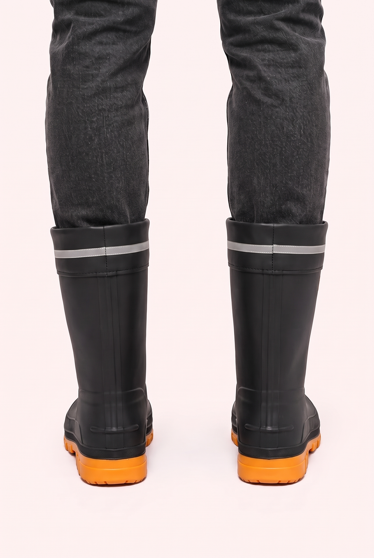 Studio photo of a person in dark grey jeans wearing black mid calf rain boots with orange lugged soles, a drawstring cuff and a reflective grey band, back view