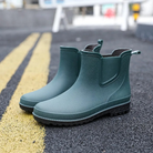 A pair of dark green rubber Chelsea style ankle boots with elastic side panels, pull tabs and black soles, standing on an asphalt road beside yellow lane markings.