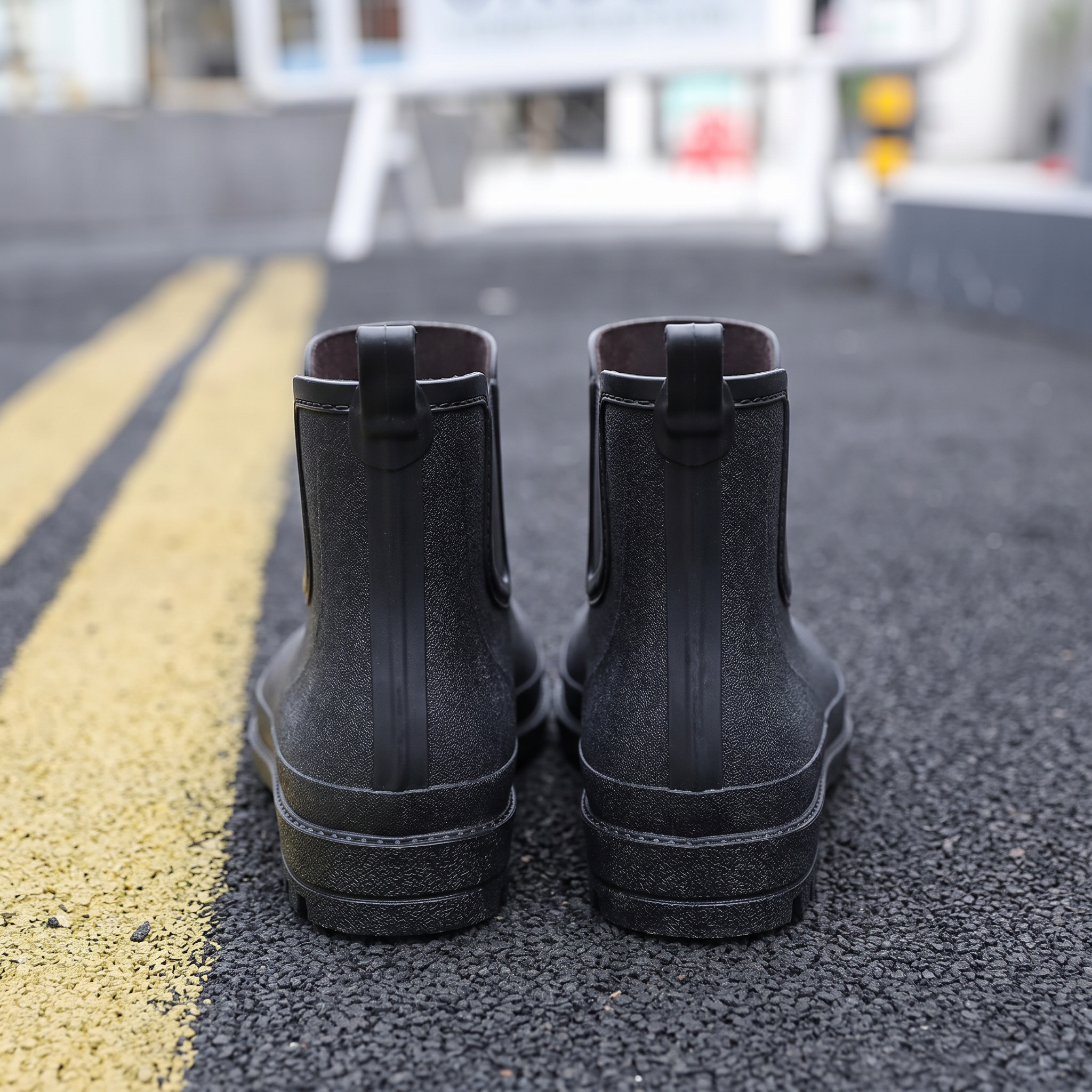 A pair of black rubber Chelsea style rain boots with elastic side panels and pull tabs standing on a street with yellow road markings, back view