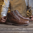 Brown faux leather lace-up ankle boots on wooden surface, person holding them | Chaps London
