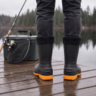 Lifestyle photo of a person standing on a wet wooden dock by a lake wearing black insulated rain boots with fleece cuffs and orange soles, with a fishing rod, landing net and tackle box beside them, back view