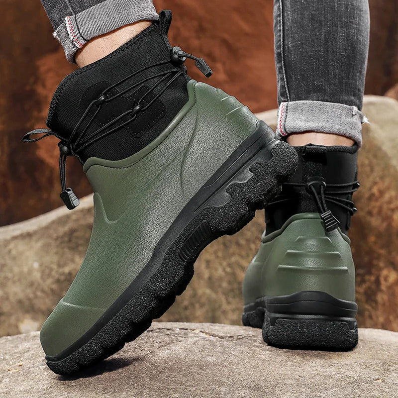 A person in rolled grey jeans stands on rocks outdoors wearing a pair of olive green rubber ankle boots with black neoprene cuffs, elastic toggle laces and thick black lug soles.