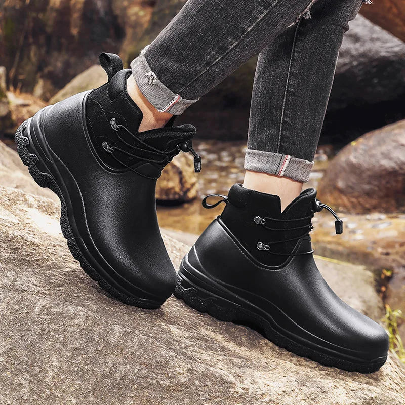 A person in cuffed grey jeans wears a pair of black ankle rain boots with a suede-look collar and bungee lacing, standing on rocks outdoors beside a stream.