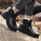A person in cuffed grey jeans wears a pair of black ankle rain boots with a suede-look collar and bungee lacing, standing on rocks outdoors beside a stream.