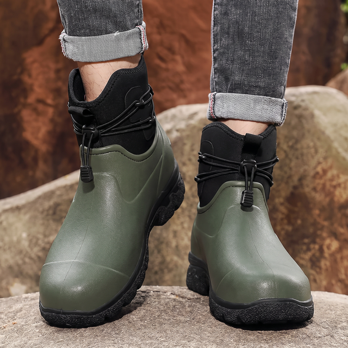 A person in rolled grey jeans stands on rocks outdoors wearing a pair of olive green rubber ankle boots with black neoprene cuffs, elastic toggle laces and thick black lug soles, front view