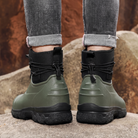 A person in rolled grey jeans stands on rocks outdoors wearing a pair of olive green rubber ankle boots with black neoprene cuffs, elastic toggle laces and thick black lug soles, back view