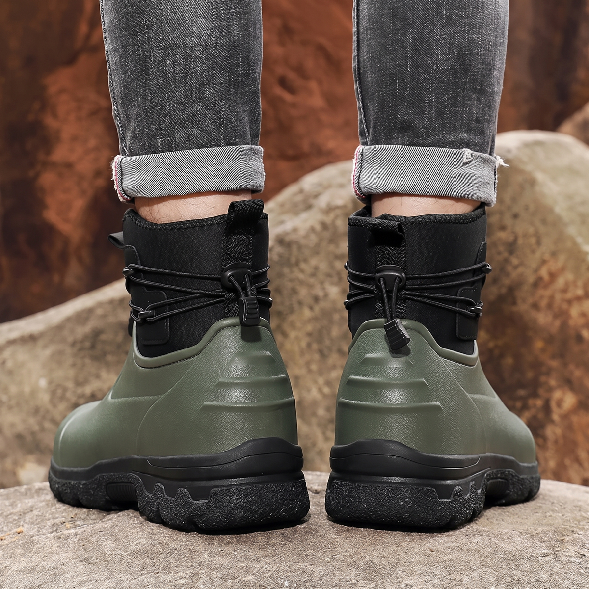 A person in rolled grey jeans stands on rocks outdoors wearing a pair of olive green rubber ankle boots with black neoprene cuffs, elastic toggle laces and thick black lug soles, back view