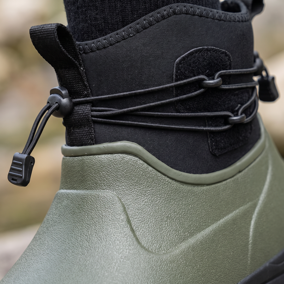Olive green rubber ankle rain boot with a black neoprene collar, bungee drawstring lacing and a black lugged sole, shown in three quarter view on a white background, close-up view