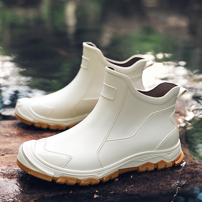 A pair of white ankle-high rubber boots with tan soles displayed outdoors on a wet rock near water.