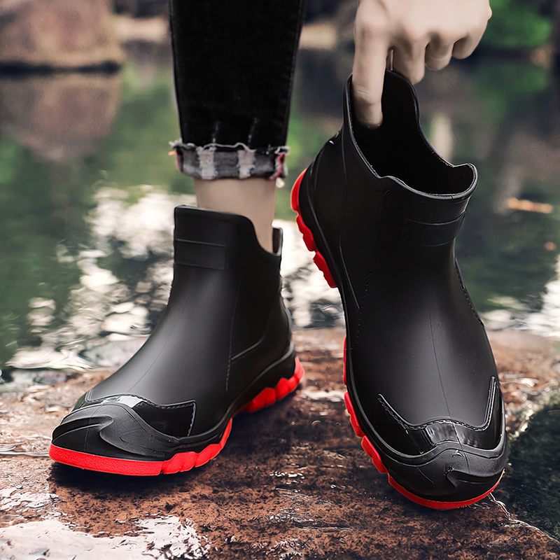 A pair of black ankle rain boots with red soles, one worn on a foot and one held by a hand, photographed outdoors near water.