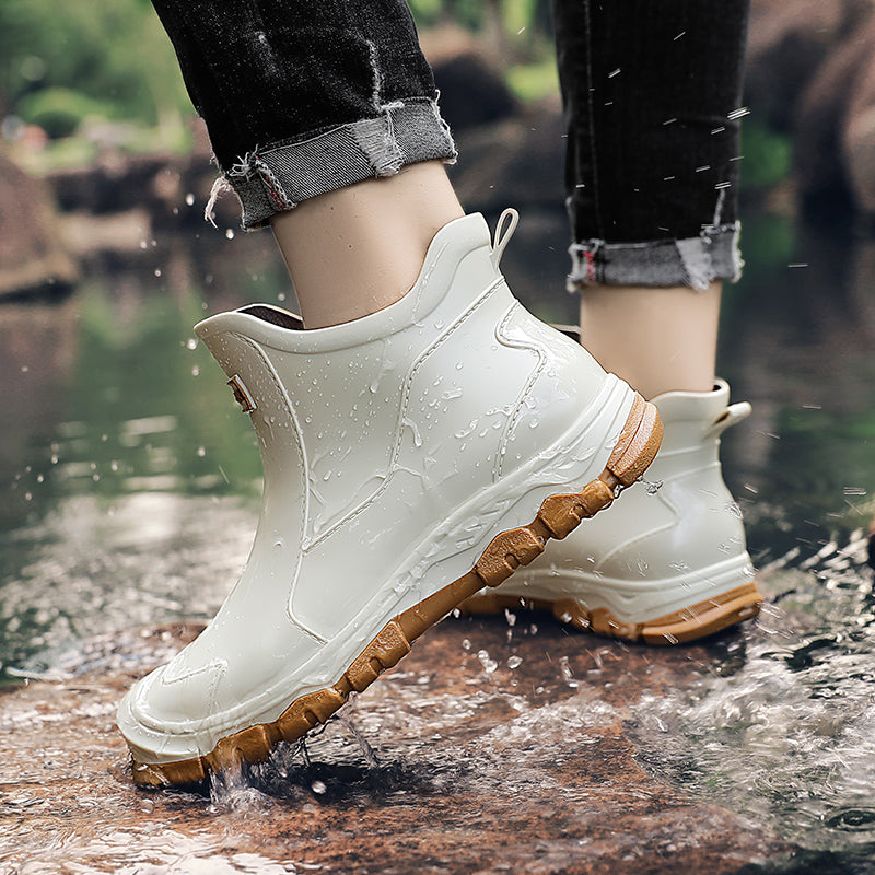 A person wearing cream colored ankle rain boots with tan textured soles stands on wet rocks near water